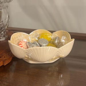 May include: A cream-colored ceramic bowl shaped like a scalloped seashell with an iridescent finish. The bowl contains a mix of wrapped candies, including silver-foiled chocolate drops, a red and white striped peppermint, and yellow candies.