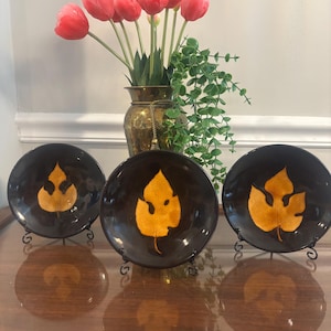 May include: Three circular black ceramic bowls, each featuring a single embossed golden-yellow leaf design in the center. The bowls are displayed on small black metal stands, arranged in a row in front of a brass vase filled with pink tulips.