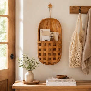 Può includere: Un organizer da parete ovale in legno intrecciato con un dettaglio a forma di stella sulla parte superiore. Il cestino contiene diverse buste e un portachiavi in metallo. È montato su una parete sopra una console in legno con un vaso e libri.