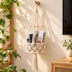 May include: A cream-colored macrame hanging basket featuring intricate knotted patterns, wooden beads, and a long tassel at the bottom. The basket holds a magazine and two remote controls, suspended from a wooden ring on a wall hook.