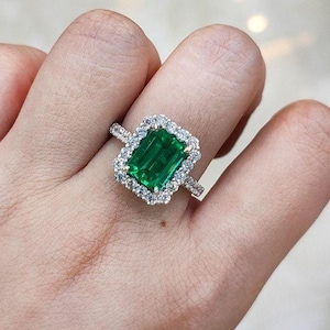 May include: A ring featuring a large, rectangular emerald-cut green gemstone at the center, surrounded by a halo of small, round, clear white diamonds. The band is made of a metallic material and is also set with small, clear white diamonds.