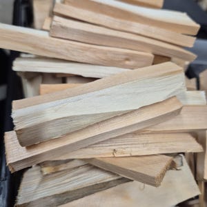 Op de afbeelding: Een verzameling lichtgekleurde, rechthoekige houten aanmaakhoutjes gestapeld in een hoop. Elk stuk hout heeft een natuurlijk nerfpatroon met verschillende tinten beige en crème, met ruw gesneden randen en een droge, vezelige textuur.