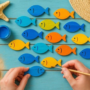 May include: A collection of small wooden fish cutouts painted in bright blue, yellow, and orange, arranged on a blue wooden surface with a starfish, a straw hat, a small container of blue paint, and smooth white pebbles nearby.