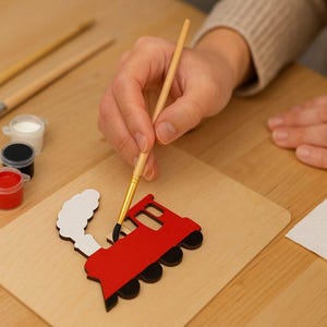 Peut inclure: Une découpe de train en bois en cours de peinture avec un pinceau fin. Le train est rouge avec un panache de fumée blanc. À proximité se trouvent deux autres pinceaux, trois petits pots en plastique contenant de la peinture rouge, noire et blanche.