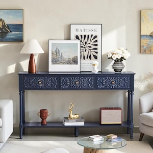May include: A navy blue console table with three drawers featuring intricate floral carvings and small gold knobs. The table has four slender legs and a lower shelf holding a brown vase, a gold deer figurine, books, and a rectangular speaker.