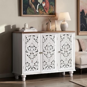 May include: A white wooden sideboard featuring three doors with intricate, carved floral patterns and small round knobs. The cabinet has a natural wood-toned top surface and rests on four rounded feet, providing a sturdy base for the storage unit.