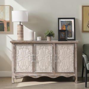 May include: A rectangular wooden sideboard featuring three doors with embossed off-white floral panels. The cabinet has a weathered light brown finish, a scalloped bottom edge, and vertical metal handles on each of the three cabinet doors.