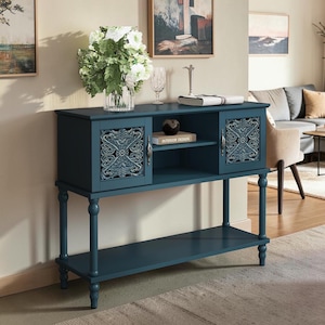 May include: A dark teal console table featuring two cabinets with intricate carved floral patterns on the doors, a central open shelf, and a lower storage shelf. The piece stands on four turned legs and includes silver-toned metal hardware.