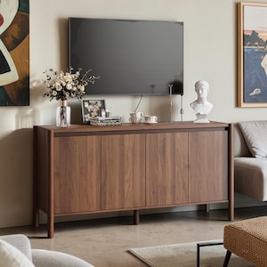 May include: A rectangular wooden sideboard with four cabinet doors featuring a dark brown wood grain finish. The unit stands on four cylindrical legs and is topped with a clear glass vase of dried flowers, books, two white cups, and a white bust.