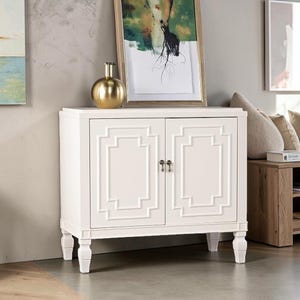 May include: A white wooden cabinet featuring two doors with a raised geometric rectangular frame design. The cabinet stands on four carved legs and includes two small metallic knobs for opening the doors. The finish is a solid matte white.