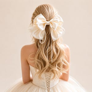 Puede incluir: Un lazo grande para el pelo de satén color crema con múltiples bucles y cintas largas y finas unidas al centro. El lazo está sujeto en el cabello largo, ondulado y rubio, con un acabado suave y brillante y una pequeña etiqueta de marca en tono dorado.
