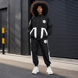 May include: A black tracksuit featuring a zip-up jacket and matching pants with white accents. The jacket has a high collar and white geometric panels at the waist. Both pieces display a circular white MCM logo patch and a subtle tonal monogram pattern.