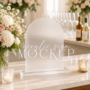May include: A clear, arched acrylic sign with a rectangular base, featuring the text "acrylic sign MOCKUP" in white script and serif fonts. The sign is positioned upright on a marble surface, displaying a smooth, transparent surface finish.