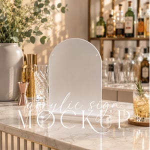 May include: A blank, white, arched acrylic sign with two small holes at the top, standing upright on a clear rectangular base. The sign features the text "acrylic sign MOCKUP" overlaid in white script and bold sans-serif lettering.