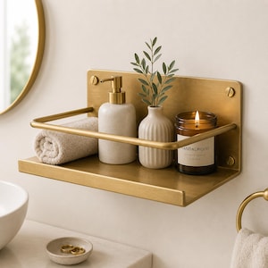Puede incluir: Un estante rectangular de pared hecho de latón con una barra protectora frontal. El estante sostiene una toalla beige enrollada, un dispensador de jabón de cerámica moteada con bomba de latón, un pequeño jarrón acanalado y una vela Sandalwood.