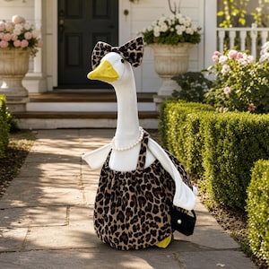 Puede incluir: Una figura de ganso blanco que lleva un lazo con estampado de leopardo en la cabeza, un collar de perlas blancas y un vestido con estampado de leopardo con mangas blancas. El ganso tiene un pico amarillo y lleva un bolso negro con una perla.