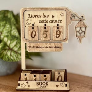 May include: A light-colored wooden book tracker display featuring the text "Livres lus cette année", "Bibliothèque de Sandrine", and "BOOK LOVER". It includes three hanging number tiles showing 059, a decorative lantern cutout, and a base with extra number tiles.