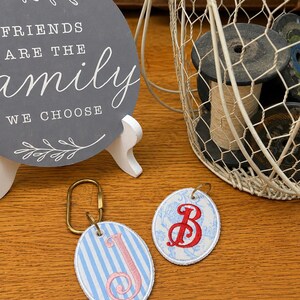 May include: Two oval-shaped fabric keychains with embroidered letters. One features a light blue and white striped pattern with a pink letter J, and the other has a light blue floral pattern with a red letter B. Both include metal rings.