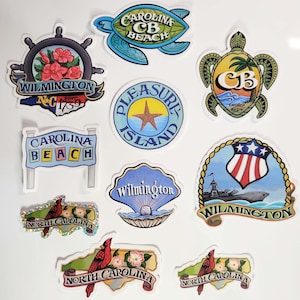 May include: Assortment of ten colorful stickers featuring various designs related to Wilmington, North Carolina. The stickers feature images of sea turtles, a lighthouse, a ship, a seashell, a star, and a cardinal. The stickers also include text such as "Carolina Beach", "Wilmington", "North Carolina", and "Pleasure Island".