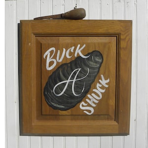 May include: A wooden sign with a painted image of an oyster and the text "Buck A Shuck" in white paint.