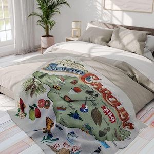 May include: A beige throw blanket with a colorful illustration of the state of North Carolina. The illustration includes various icons representing different cities and landmarks, such as the state capitol building, a basketball hoop, a donut, and a lighthouse. The text "A you're in North Carolina" is written at the top of the illustration.