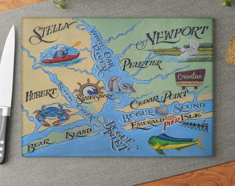 May include: Rectangular cutting board with a colorful map design. The map features coastal areas with illustrations of a crab, fish, dolphins, and a kayaker. Text includes place names like Stella, Newport, and Emerald Isle.
