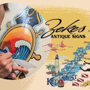 May include: A man is painting a wooden sign with a nautical theme. The sign features a ship's wheel, a wave, and a pirate hat. The sign is surrounded by a yellow background with a map of the Florida Keys and the text "Zekes Antique Signs".
