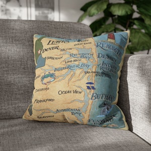 May include: A decorative throw pillow with a map of the Delaware coast. The map features the names of towns and cities along the coast, including Lewes, Dover, Rehoboth Beach, Bethany Beach, and Fenwick Island. The map is surrounded by a beige border.