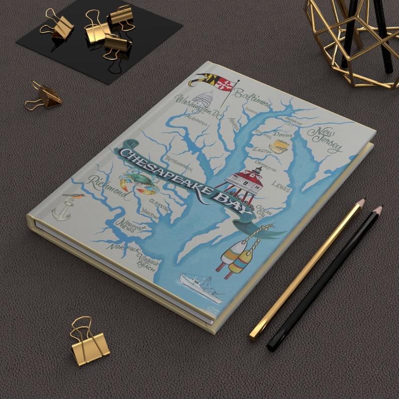 May include: A white notebook with a map of the Chesapeake Bay on the cover. The map is illustrated with blue water, green land, and red and white buoys. The title "CHESAPEAKE BAY" is written in a decorative font. The notebook is lying on a grey surface with gold and black pencils.