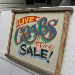 May include: A wooden sign with a hand-painted message that reads "LIVE CRABS for SALE!" in colorful lettering. The sign is framed in a rustic wooden frame.