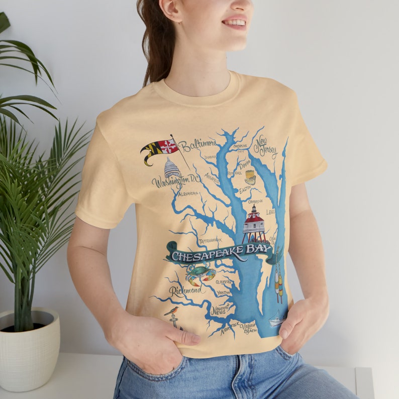 May include: A light yellow t-shirt with a map of the Chesapeake Bay printed on the front. The map includes the cities of Baltimore, Washington D.C., Richmond, and Norfolk. The shirt also features a lighthouse, crab, and other nautical imagery.
