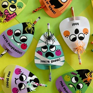 Op de afbeelding: Een verzameling Halloween-potloodtoppers in verschillende kleuren, waaronder groen, paars en oranje. Elke topper heeft een ander monsterontwerp met een potlood erin. Sommige toppers hebben tekst zoals "You're fangtastic!" en "Hey ghoul!"