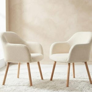 Peut inclure: Deux fauteuils dotés d'un revêtement en tissu crème à côtes verticales. Chaque chaise présente un dossier incurvé, des accoudoirs intégrés et quatre pieds fuselés en bois clair qui s'étendent vers l'extérieur depuis la base.
