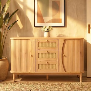 Könnte beinhalten: Ein Sideboard aus hellem Holz mit zwei Seitenschränken mit vertikalen Lamellentüren und drei mittigen Schubladen mit geflochtenen Rattaneinsätzen. Das Möbelstück steht auf vier konischen Beinen mit goldfarbenen Kappen und runden Holzknöpfen.