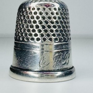 May include: A silver-colored metal thimble featuring a textured, dimpled surface on the upper portion and a smooth, polished band at the base. The lower section displays an engraved monogram with the letters E and R in a cursive script style.