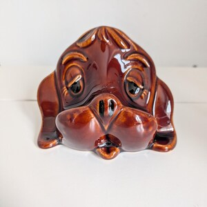 May include: A glossy, dark brown ceramic figurine shaped like the head of a dog with long, drooping ears, large eyes, and a prominent snout. The piece features a smooth, reflective glaze with darker brown shading around the facial features.