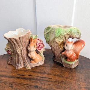 May include: Two ceramic planters shaped like tree stumps. The left planter features a light brown rabbit and pink flowers. The right planter features a brown squirrel nestled under green leaves. Both have a glossy finish and hollow centres.