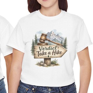 Puede incluir: Una camiseta blanca con un gráfico de un poste de madera con el texto "Verdict: Take a Hike" en negro. El letrero está rematado con un mazo de madera y se sitúa sobre un fondo de pinos verdes y picos de montañas cubiertos de nieve.