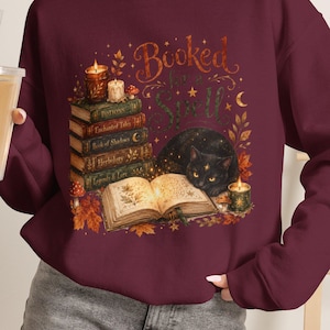 Puede incluir: Una sudadera de manga larga color granate con un estampado gráfico de un gato negro tumbado junto a un libro abierto, una pila de libros, velas y hojas de otoño. El texto "Booked for a Spell" aparece impreso en una fuente estilizada.