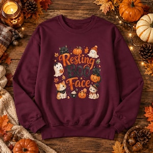 Puede incluir: Una sudadera de cuello redondo color granate con un estampado gráfico que incluye el texto Resting Boo Face, rodeado de ilustraciones de pequeños fantasmas, gatitos, calabazas, hojas de otoño y una taza de café con nata en naranja y negro.