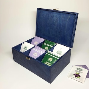 Könnte beinhalten: Eine rechteckige Teebox aus Holz mit tiefblauer Beize, einem Klappdeckel und einem Metallverschluss. Das Innere ist in sechs Fächer unterteilt, die verschiedene Teebeutel in weißer, grüner und violetter Verpackung mit Aufdruck enthalten.