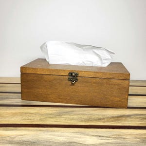 May include: A rectangular wooden tissue box holder with a medium brown stained finish. The box features a hinged lid secured by a small, antique brass-coloured metal latch centred on the front. A white tissue is protruding from the top opening.