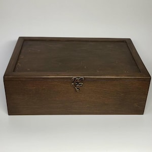 Pode incluir: Uma caixa retangular de madeira com acabamento castanho escuro e tampa plana e lisa. A caixa apresenta um fecho metálico de cor latão em estilo antigo, centrado no painel frontal, usado para prender a tampa à base do recipiente.