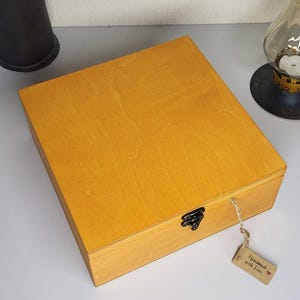 May include: A square wooden box with a light brown finish and a visible wood grain pattern. The box features a dark metal latch on the front edge and a small rectangular paper tag attached by a string, which reads Handmade with Love.