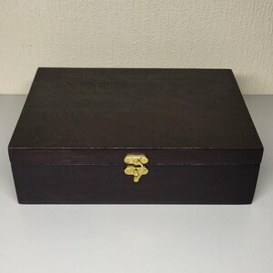 Pode incluir: Uma caixa retangular de madeira com acabamento em marrom escuro e um padrão de veios de madeira visível. A caixa apresenta um fecho de metal dourado centralizado no painel frontal, usado para prender a tampa à base do recipiente.