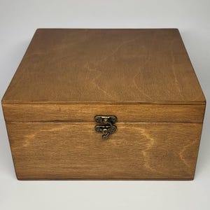 Puede incluir: Una caja cuadrada de madera con un acabado marrón medio y patrones de veta de madera visibles. La caja cuenta con una tapa con bisagras y un mecanismo de cierre central de metal color latón antiguo asegurado con tornillos.