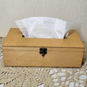 May include: A rectangular light brown wooden tissue box holder with a hinged lid and a black metal latch on the front. A stack of white paper tissues is protruding from the oval-shaped opening on the top surface of the wooden container.