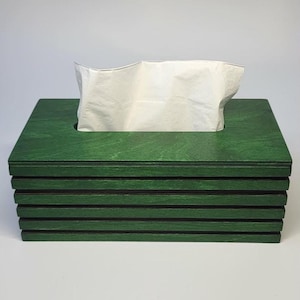 Könnte beinhalten: Ein rechteckiger Taschentuchbox-Überzug aus grün gebeiztem Holz mit geschichtetem Lamellendesign. Die Box verfügt über eine horizontale Öffnung auf der Oberseite, durch die ein einzelnes weißes Papiertaschentuch nach oben herausragt.