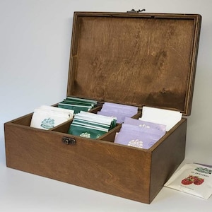 Könnte beinhalten: Eine rechteckige Teebox aus Holz mit Klappdeckel und Metallverschluss, die drei Innenfächer mit verschiedenen Teebeuteln in weißer, grüner und hellvioletter Verpackung enthält, wobei ein zusätzlicher Teebeutel außerhalb der Box liegt.
