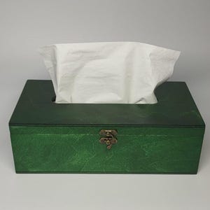 May include: A rectangular tissue box cover with a dark green wood grain finish. The box features a hinged lid with a metallic bronze-colored latch on the front. A single white paper tissue is pulled through the rectangular opening on the top.